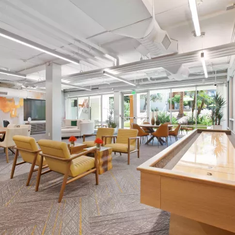 Vibrant resident lounge with a shuffleboard table, colorful accent wall, modern seating areas, and floor-to-ceiling windows that open to a lush outdoor courtyard.