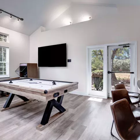 Modern resident lounge with a rustic-style air hockey table, mounted flat-screen TV, shuffleboard, bar seating, and large windows providing natural light.