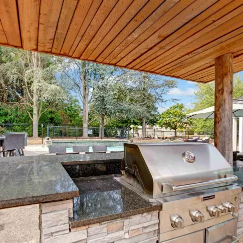 Outdoor grilling station with a built-in stainless steel KitchenAid grill, granite countertops, and a stone base under a wooden pergola. In the background, there is a pool area, patio seating with wicker furniture, and lush trees providing a serene setting.