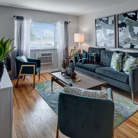 A stylish and cozy living room at Alcove at Seahurst, featuring a soft blue sofa adorned with decorative pillows, a rustic wooden coffee table with a floral centerpiece, and two modern accent chairs with gold metal frames. The space is illuminated by warm table lamps, and abstract wall art enhances the contemporary decor. An open doorway leads to a small dining area, and large windows bring in natural light, creating an inviting atmosphere. ​​