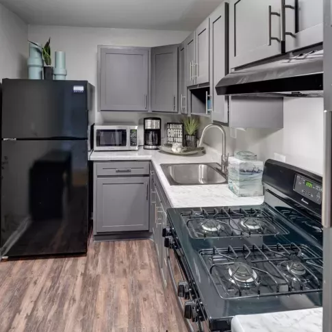 A sleek and functional kitchen at East Broad Plaza featuring gray cabinetry with brushed nickel hardware, white marble-style countertops, and black appliances, including a refrigerator, gas stove, and range hood. The compact kitchen layout is efficiently designed with a deep stainless steel sink and modern fixtures. Decorative elements like a coffee sign, potted plant, and accent jars add warmth and character to the space. ​​