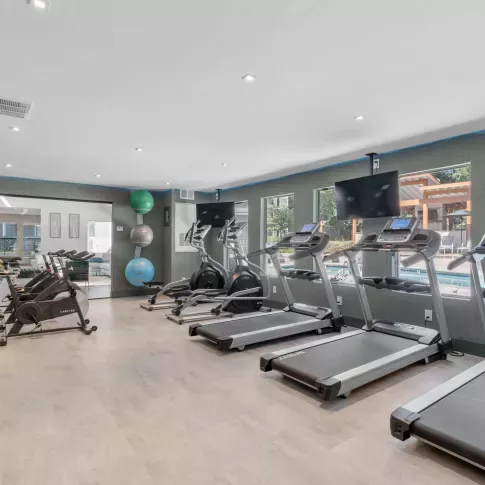 Modern fitness center with treadmills, ellipticals, stationary bikes, and free weights, featuring large windows overlooking the pool and outdoor lounge area.