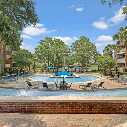 Resort-style swimming pool area surrounded by lounge chairs, palm trees, and red brick apartment buildings under a bright blue sky.