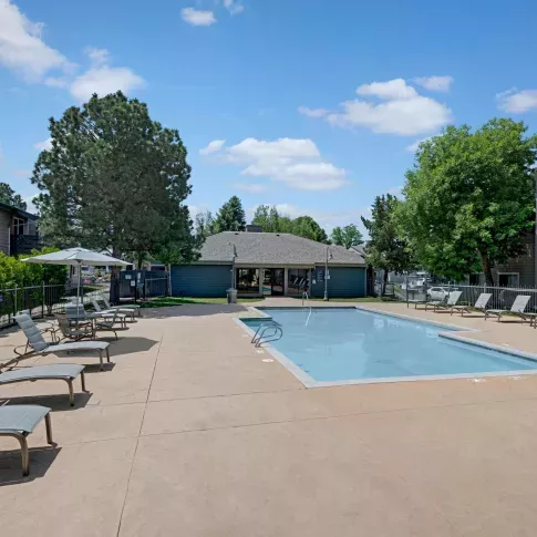 Outdoor swimming pool with surrounding lounge chairs, umbrellas, and fenced-in area, set in a residential apartment community with trees and blue skies.