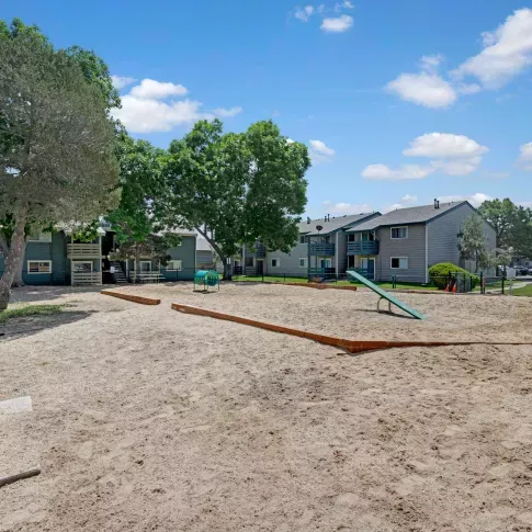Outdoor dog park with sand surface, seesaw, and play tunnel, surrounded by mature trees and apartment buildings in the background.