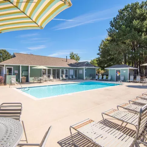 Outdoor swimming pool area with lounge chairs, patio tables, and umbrellas, surrounded by tall pine trees and a clubhouse building.