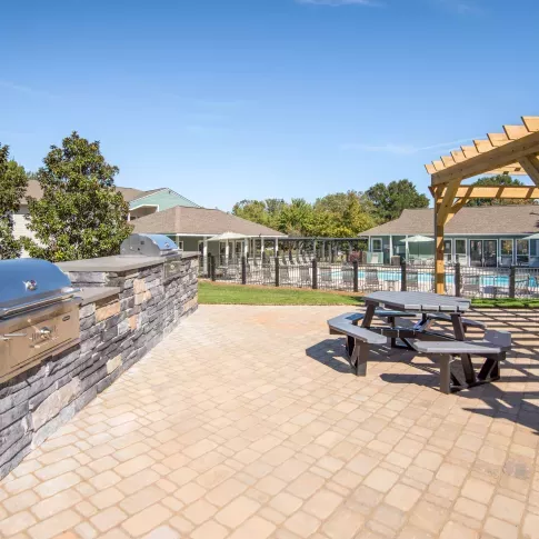Outdoor grilling area with built-in stainless steel grills, stone countertops, picnic seating under a wooden pergola, and a view of the swimming pool and clubhouse in the background.