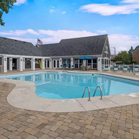 Inviting outdoor pool area with a curved swimming pool, cushioned lounge chairs, and a classic clubhouse building featuring white trim, gabled roofing, and a shaded patio with seating.