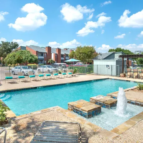 Outdoor swimming pool with stone water fountain feature, surrounded by lounge chairs with teal cushions, lush landscaping, and red brick apartment buildings in the background.