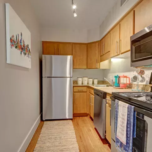 Contemporary galley kitchen with light wood cabinetry, stainless steel appliances, granite countertops, and a cozy decorative rug on wood-style flooring.