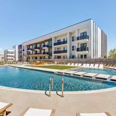Resort-style swimming pool at Mezzo Apartments, featuring a modern curved design with in-pool lounge seating and stepping stones leading to a sun deck. Surrounding the pool are white cushioned lounge chairs, shaded pergolas, and a contemporary three-story apartment building with private balconies. The outdoor area is landscaped with greenery, creating a stylish and inviting atmosphere under a bright blue sky.