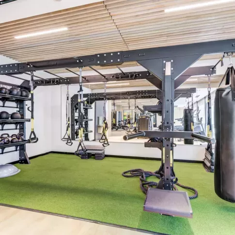 Modern fitness center at Mezzo Apartments featuring a functional training station with TRX suspension straps, kettlebells, medicine balls, a punching bag, and battle ropes. The space has a sleek design with a mirrored wall, wood-accented ceiling, and artificial turf flooring for dynamic workouts. The gym is well-lit and designed for strength training, agility, and high-intensity workouts.