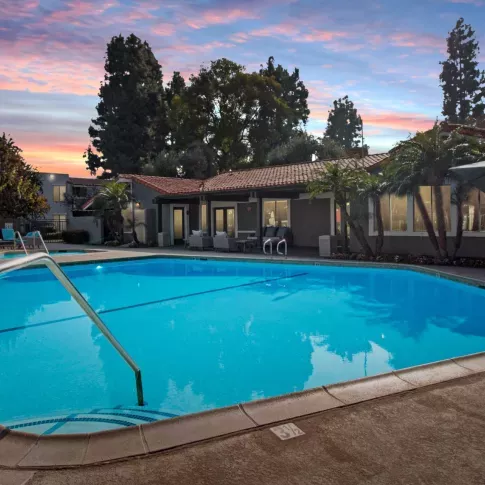 An inviting outdoor pool area at Olive Ridge Apartments, illuminated by the soft glow of the twilight sky. The pool is surrounded by tropical landscaping, including tall palm trees and lush greenery. Lounge chairs and shaded seating areas provide a relaxing environment, with the apartment community's clubhouse in the background.