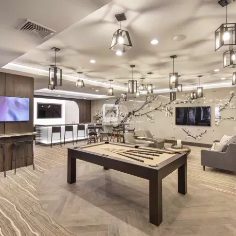 Upscale resident lounge featuring a modern pool table in the foreground, surrounded by plush seating and elegant decor. The space is illuminated by a grid of geometric pendant lights, and the back wall showcases a striking floral art installation beneath a mounted TV. A sleek bar area with high-top chairs and underlighting sits in the background, providing a luxurious and social atmosphere ideal for relaxing or entertaining.