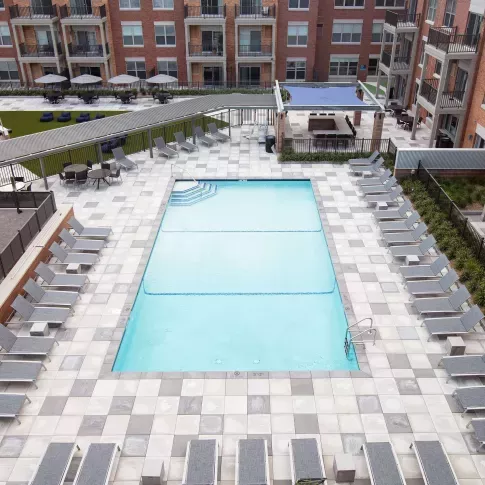 Bright and inviting courtyard pool deck featuring a rectangular swimming pool surrounded by uniform gray tile and sleek lounge chairs neatly arranged on all sides. In the background, a beautifully maintained lawn and shaded cabana area provide extra space for relaxation and social gatherings. The surrounding red-brick apartment buildings with balconies add a warm, urban atmosphere to this serene outdoor amenity space.