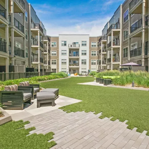 Ground-level view of a vibrant apartment courtyard showcasing artificial turf, modern outdoor seating with cushioned chairs, and well-maintained landscaping. The space is enclosed by four-story residential buildings with balconies, creating a cozy and community-focused atmosphere. The open layout and bright sky provide a welcoming outdoor area perfect for relaxation or social gatherings.