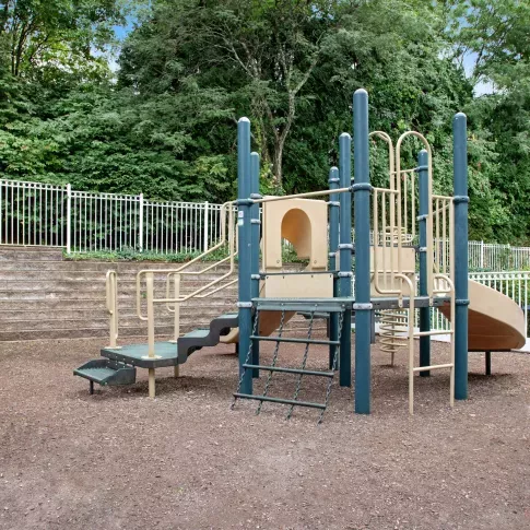 Fenced playground with slides, climbing structures, and mulch ground cover, set against terraced stone steps and tall trees.