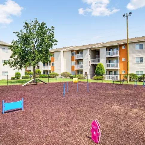 Fenced-in dog park at The Samuel with agility equipment and nearby apartment buildings in the background