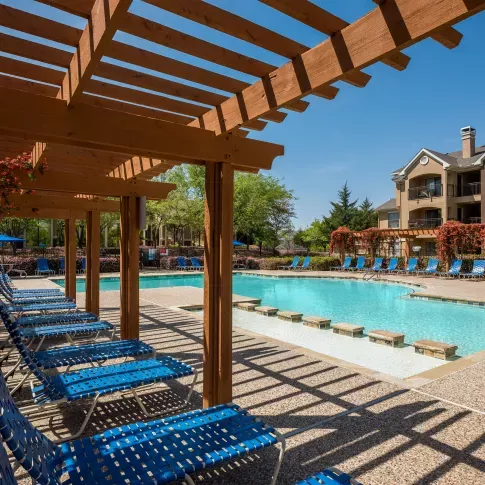 Resort-style swimming pool with stepping stones, surrounded by blue lounge chairs and wooden pergolas draped in vibrant flowering vines, adjacent to a multi-story apartment building.