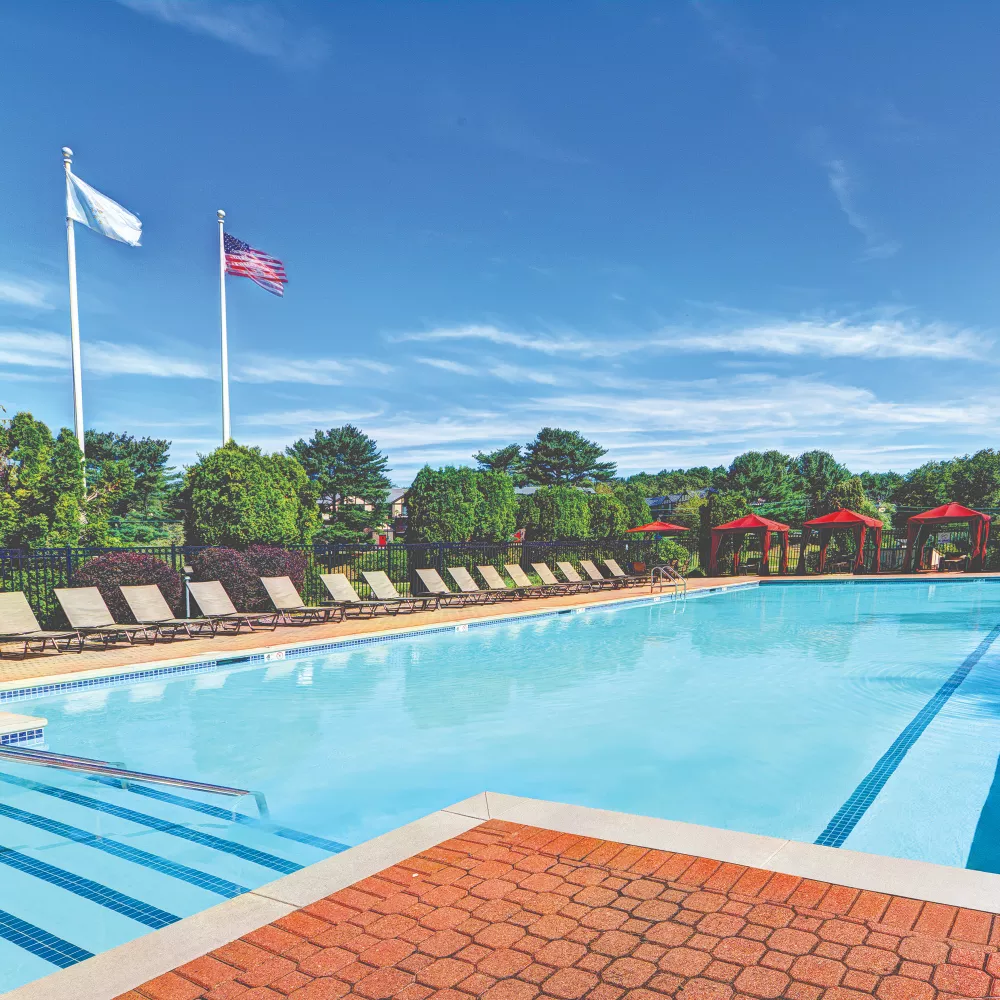 Outdoor pool with shallow step entry, lap lane, and flags flying near cabanas and loungers at Royal Crest Estates Warwick in Warwick, RI.