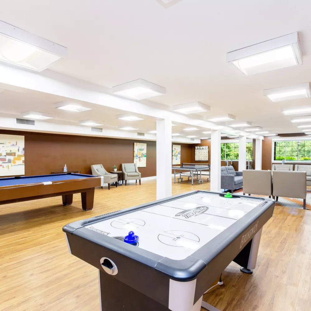 Recreation room with air-hockey and pool tables, ping-pong, and multiple seating zones near a large window at Royal Crest Estates Warwick in Warwick, RI.