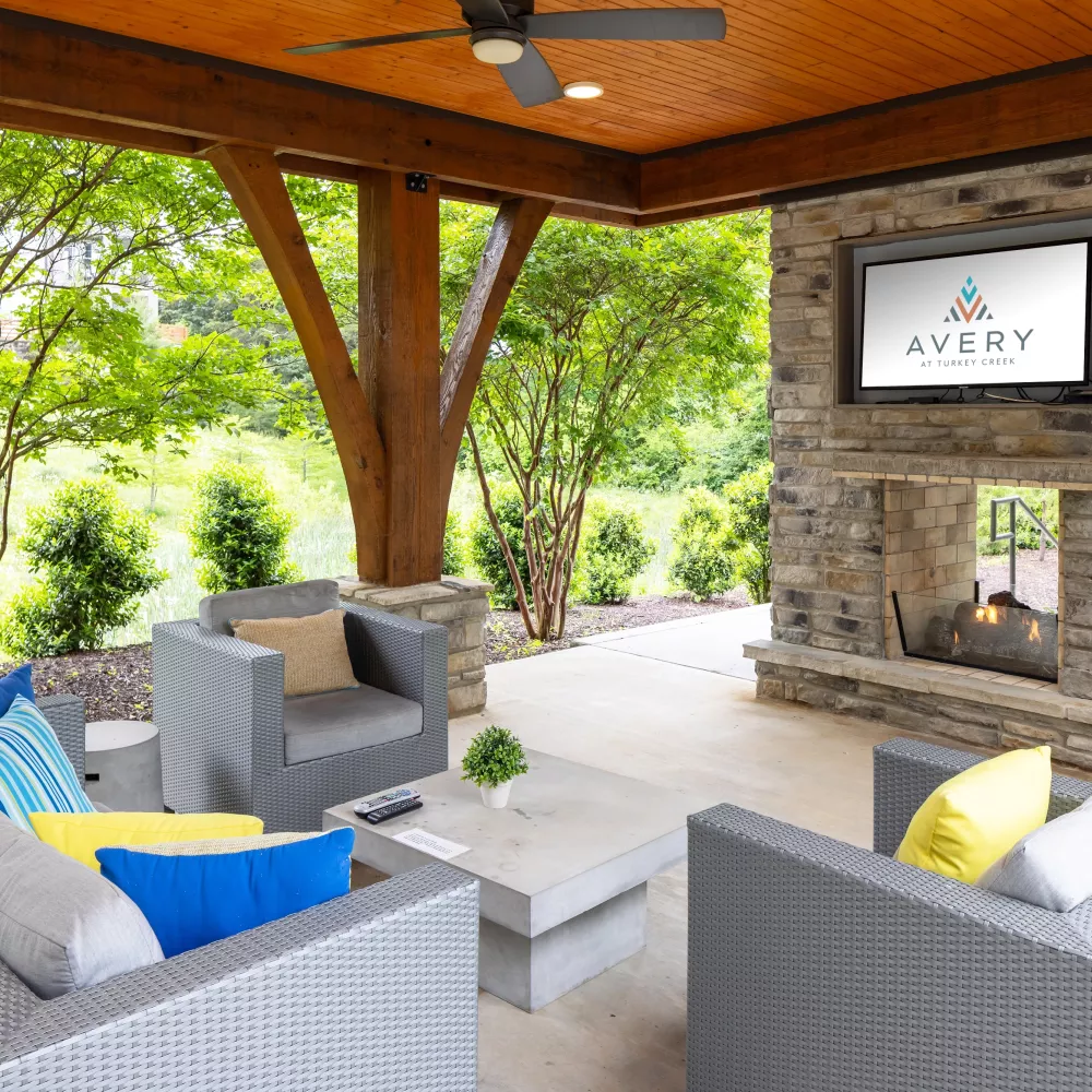 Covered outdoor lounge featuring modern wicker seating, colorful accent pillows, a stone fireplace with mounted TV, and views of landscaped greenery.