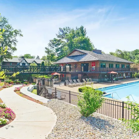 Community clubhouse and outdoor swimming pool surrounded by lounge chairs, red umbrellas, and landscaped gardens with colorful flowers, connected by a walkway and pedestrian bridge on a sunny day.