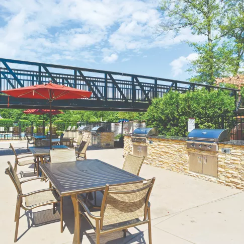 Outdoor grilling and dining area with stainless steel barbecue stations, stone countertops, tables with umbrellas, and a pedestrian bridge overhead, surrounded by greenery.