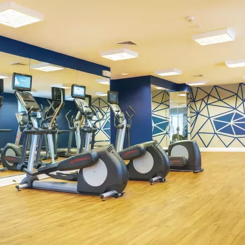 Modern fitness center with cardio machines and wall-mounted TVs, featuring mirrored walls, wood-style flooring, and bold geometric accent wall design.