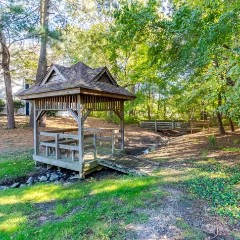 A small wooden gazebo with a shingled roof and railing stands over a narrow, rocky stream bed in a shaded, grassy area surrounded by tall pine and deciduous trees. In the background, part of a light-gray apartment building is visible among the trees, and sunlight filters through the leaves, creating a peaceful, natural setting.
