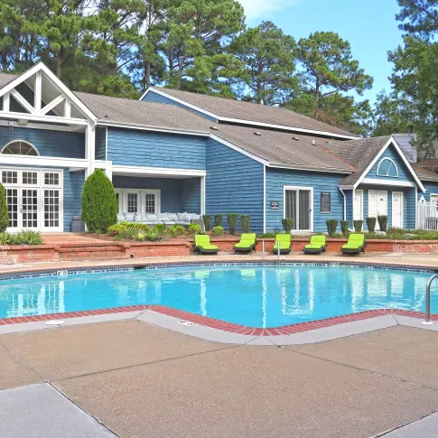 Outdoor pool area beside a blue clubhouse with white trim and a brick chimney. The pool has a curved design with red brick coping and steps leading into the water. Green lounge chairs line the pool deck, surrounded by neatly trimmed shrubs and tall trees, creating a relaxed and inviting atmosphere under a clear sky.