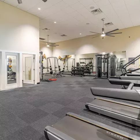 “Spacious fitness center with high ceilings, featuring treadmills, free weights, strength training machines, and large mirrors along the back wall. The room has gray carpet tiles, ceiling fans, and a separate area with stability balls and resistance equipment visible through glass doors.