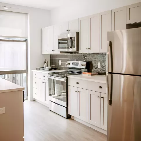Modern apartment kitchen with white shaker cabinets, stainless-steel appliances, subway-tile backsplash, and large window providing natural light.