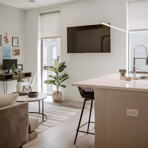 Bright modern living area with a wall-mounted TV, cozy sofa, small desk setup, large window, and a kitchen island with a barstool.