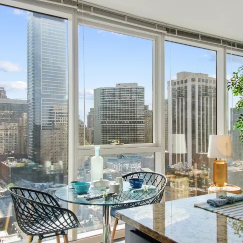 A glass dining table is positioned next to a floor-to-ceiling window offering expansive views of high-rise city buildings.