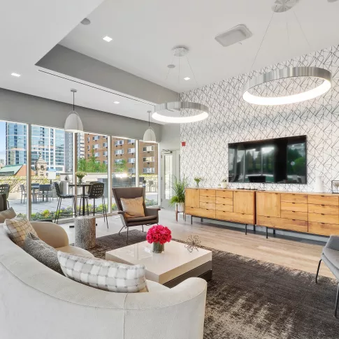 Spacious clubroom featuring a media wall with a flat-screen TV and wooden cabinetry. Large windows fill the room with natural light, highlighting the geometric accent wall and modern furniture.