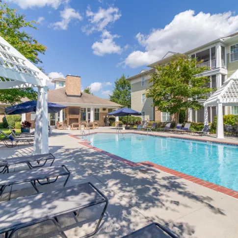 Resort-style pool area with lounge chairs, white pergolas, and a gazebo, surrounded by apartment buildings under a blue sky.