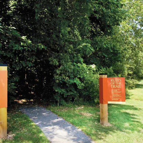 Orange signs mark the entrance to a paved nature trail winding through lush green trees and grass on a sunny day.
