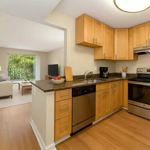Bright kitchen featuring light wood cabinets, stainless steel appliances, and a glimpse of a cozy living room.