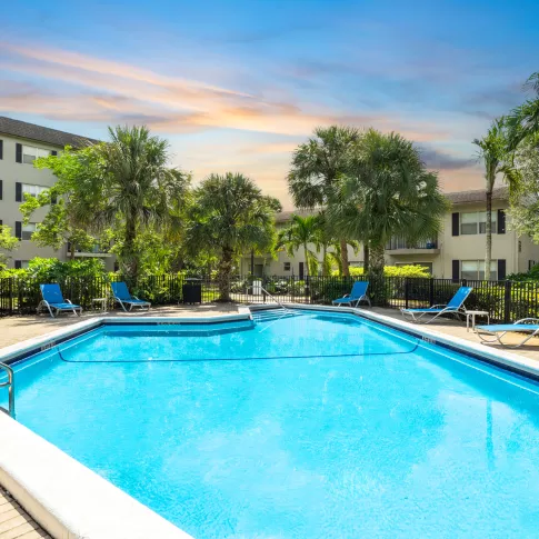 Outdoor swimming pool with blue lounge chairs, surrounded by palm trees and apartment buildings under a clear sky.