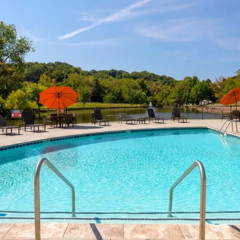 Bright blue swimming pool with lounge chairs, orange umbrellas, and a nearby pond under a clear sky.