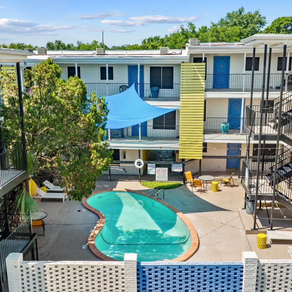 Aerial photograph of a vibrant apartment complex courtyard with a kidney-shaped pool, blue sunshade, and colorful buildings.