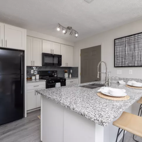 Modern kitchen with light cabinets, black appliances, granite island with two stools, and light wood flooring.