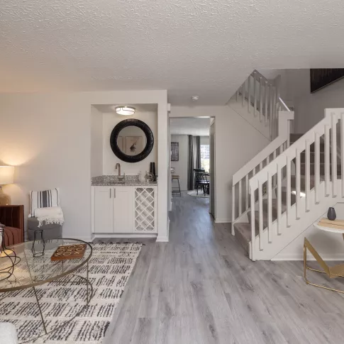 Modern living room with brown sofa, glass coffee table, wet bar, and white staircase.