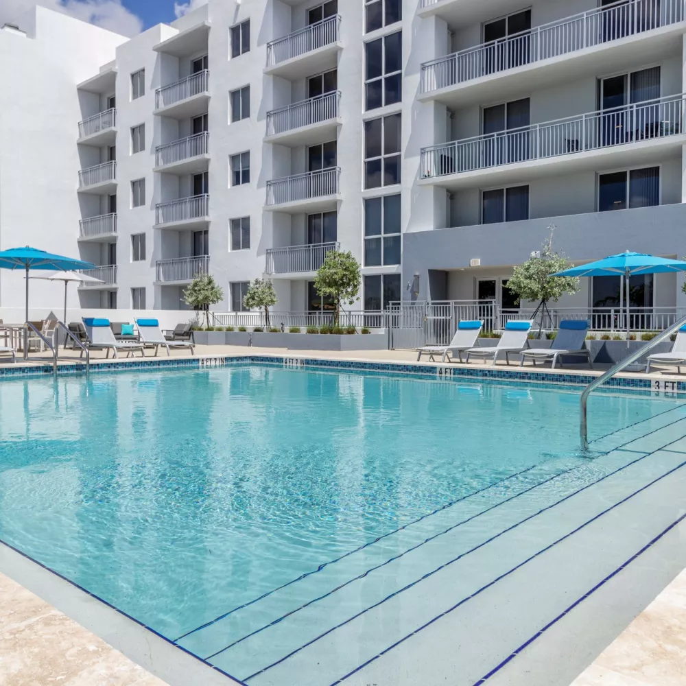 Modern apartment building with a large swimming pool, blue lounge chairs, and umbrellas under a partly cloudy sky.