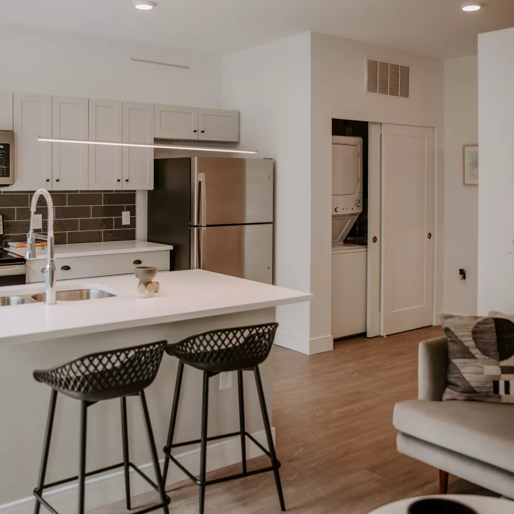 Bright, open-concept apartment featuring a modern kitchen with white cabinets, dark backsplash, island, and adjacent living space.
