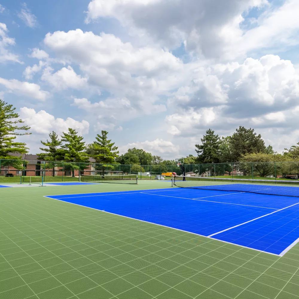 Blue tennis court on a green grid-patterned surface, framed by trees and buildings under a sunny, cloudy sky.