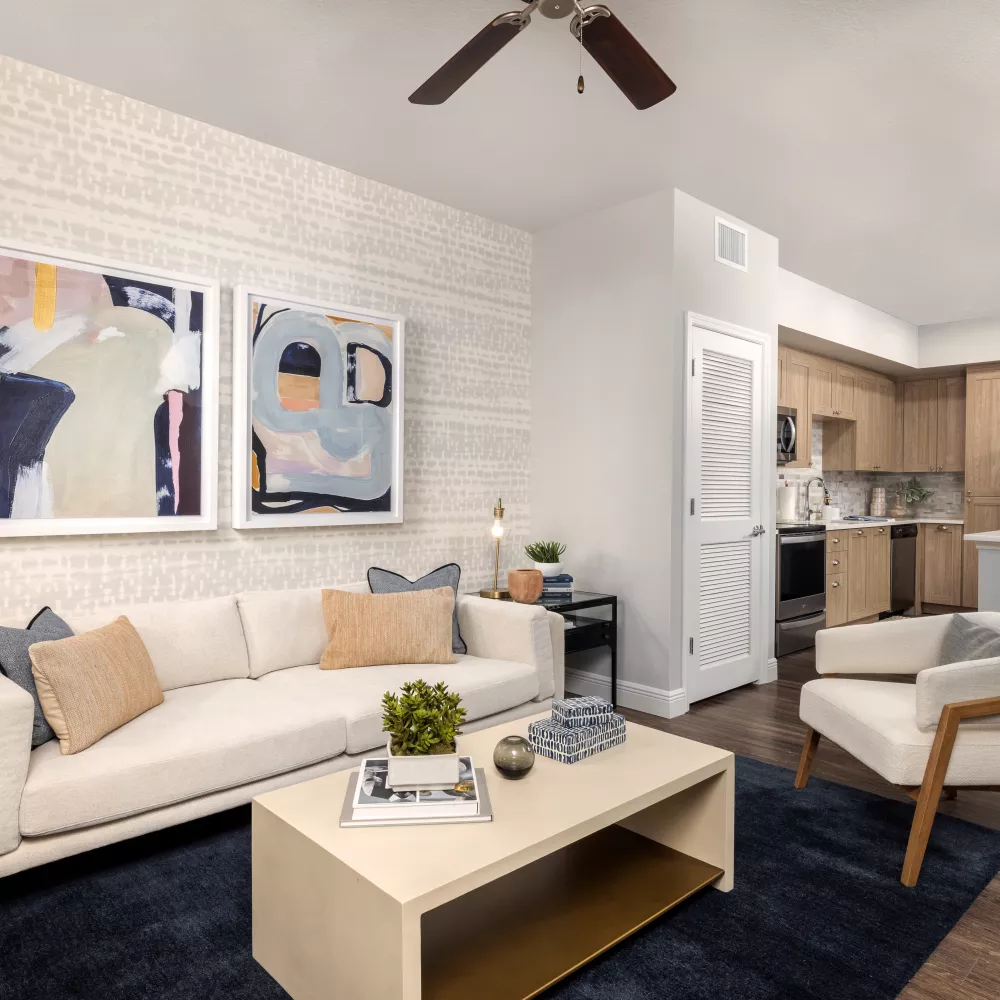 Well-lit living room with cream sofa, abstract art, and dark blue rug, open to a modern kitchen with light wood cabinets.