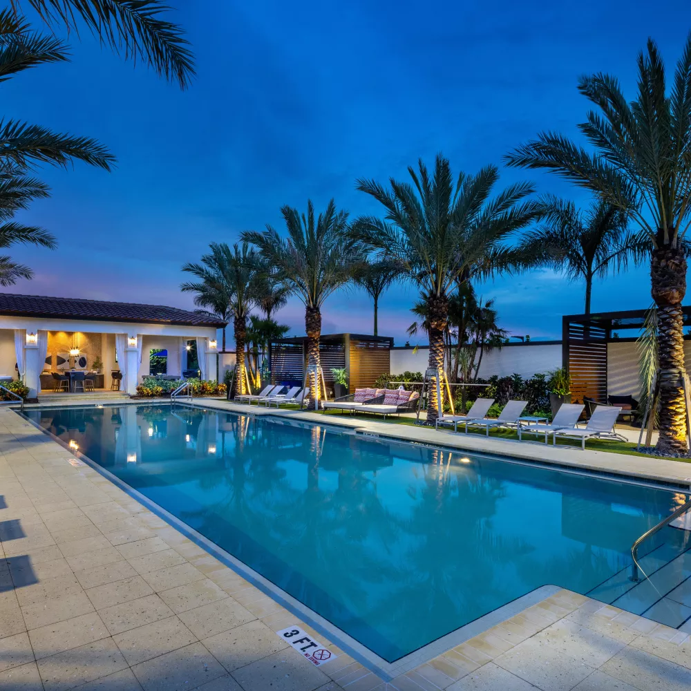 Tropical resort-style pool with palm trees, lounge chairs, and a lit cabana at twilight.