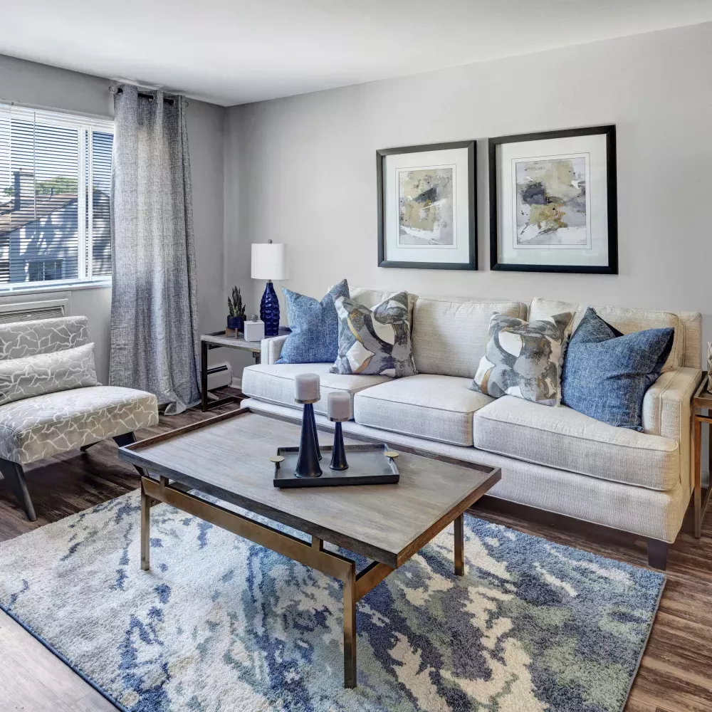 Inviting living room with light couch, blue pillows, patterned armchair, wood coffee table, blue abstract rug, and window.